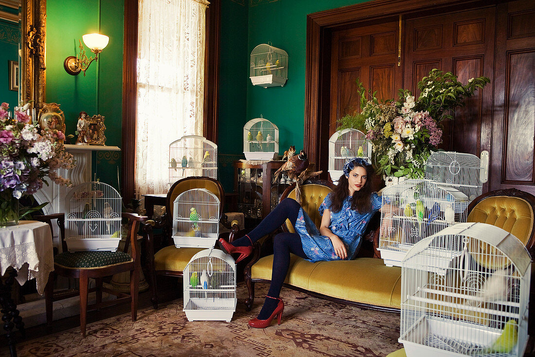 A dark-haired woman sitting on a sofa in a living room surrounded by birdcages