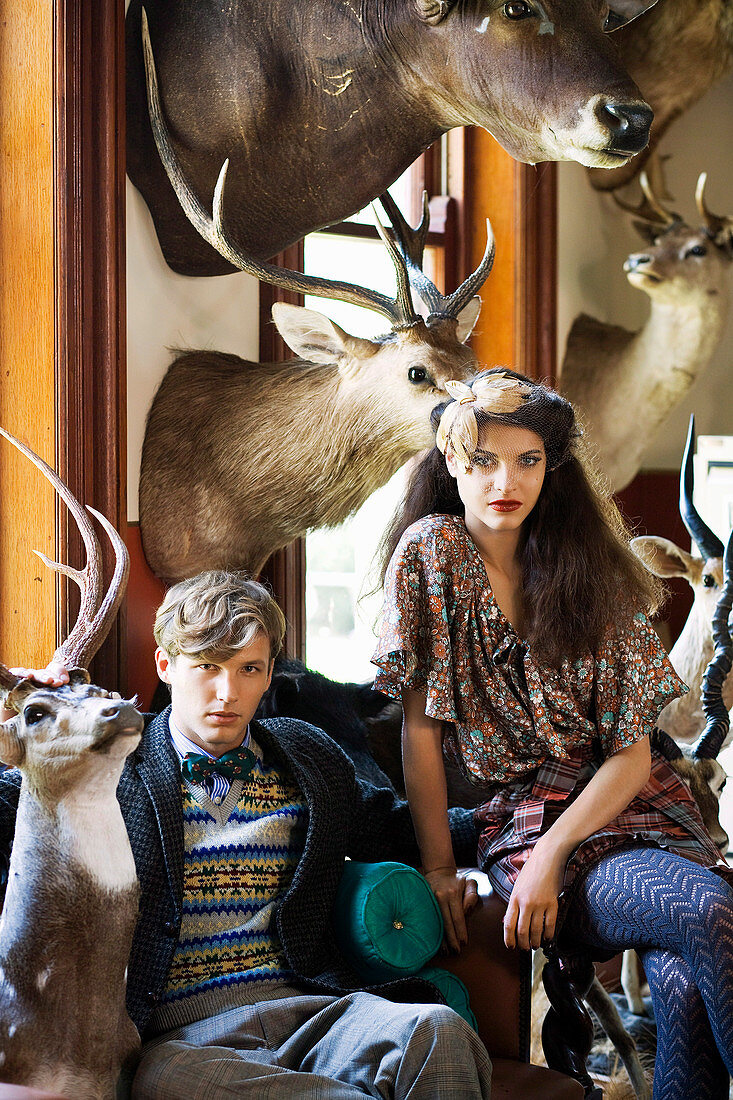 A dark-haired woman wearing a headpiece and a man sitting in a lounge surrounded by hunting trophies