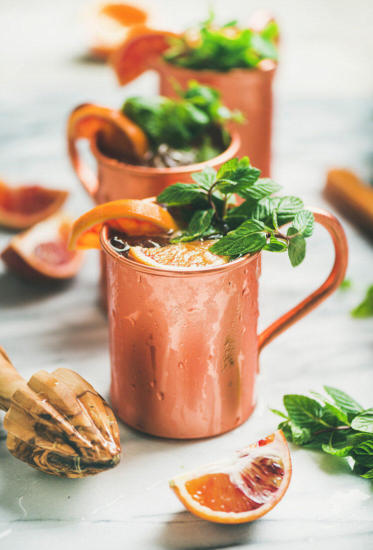 Blood orange Moscow mule alcohol cocktails with fresh mint leaves and ice in copper mugs over white marble background