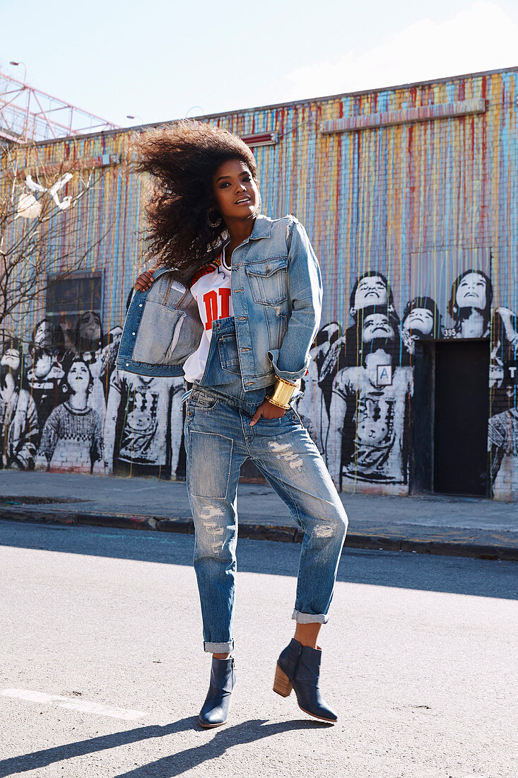 A dark-haired woman wearing denim dungarees and a denim jacket standing against a graffitied wall