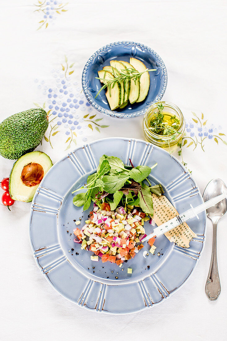 A syringe on a plate on avocado and tomato salad (seen from above)