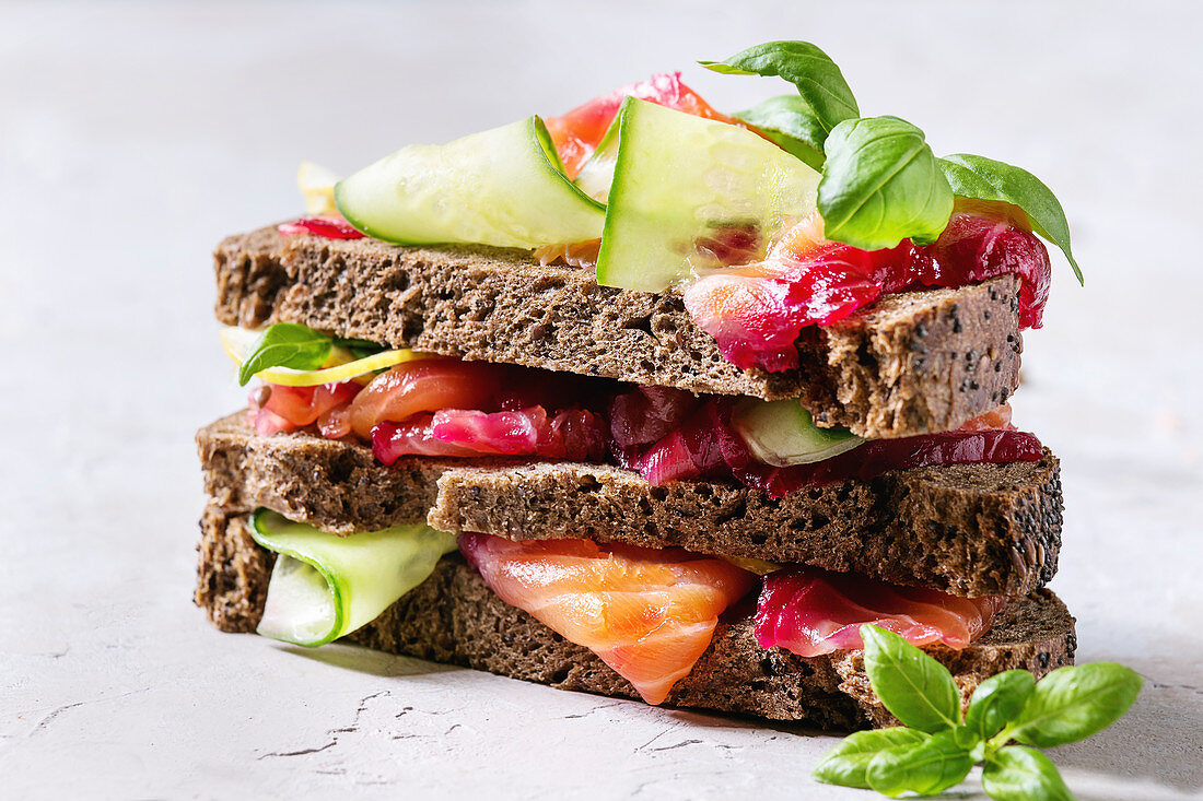 Sliced beetroot marinated salmon sandwiches with rye bread, cucumber, basil and lemon served in stack on wooden cutting board
