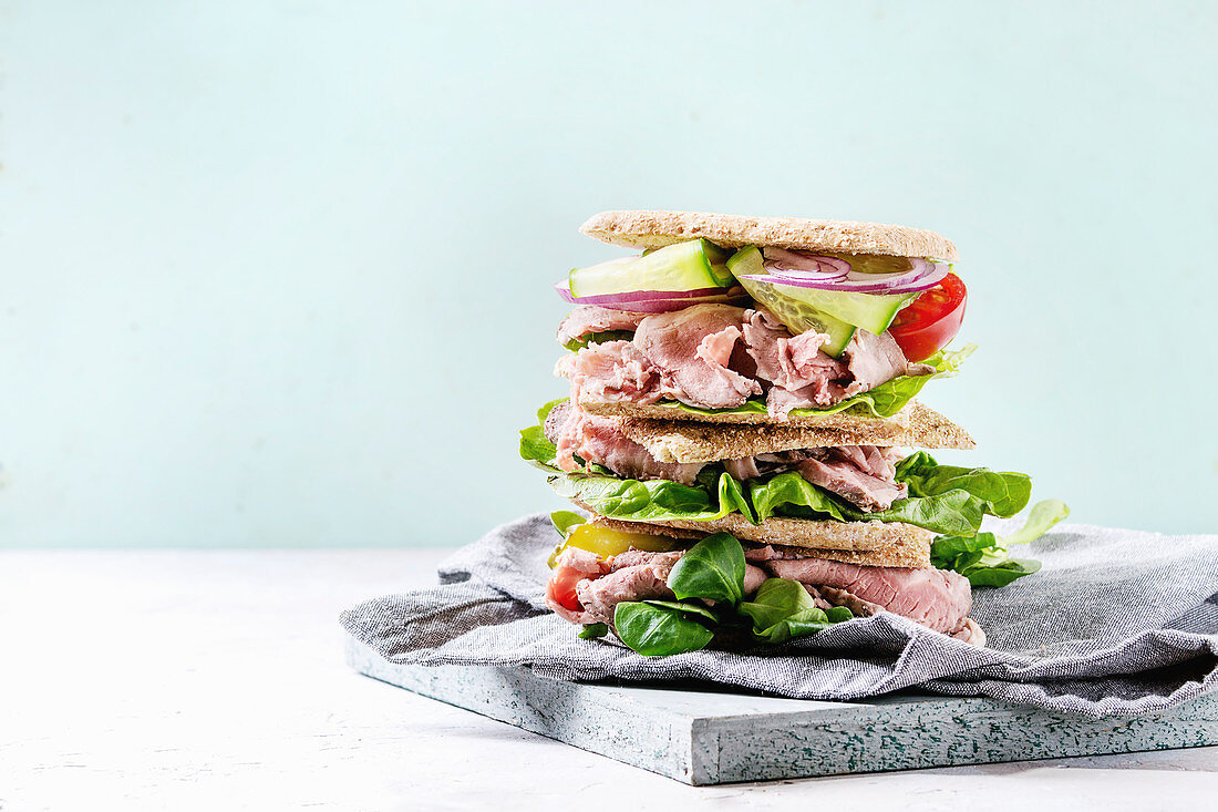 Beef and vegetables sandwiches with sliced meat, cucumber, green salad, rye whole grain bread in stack on wooden tray