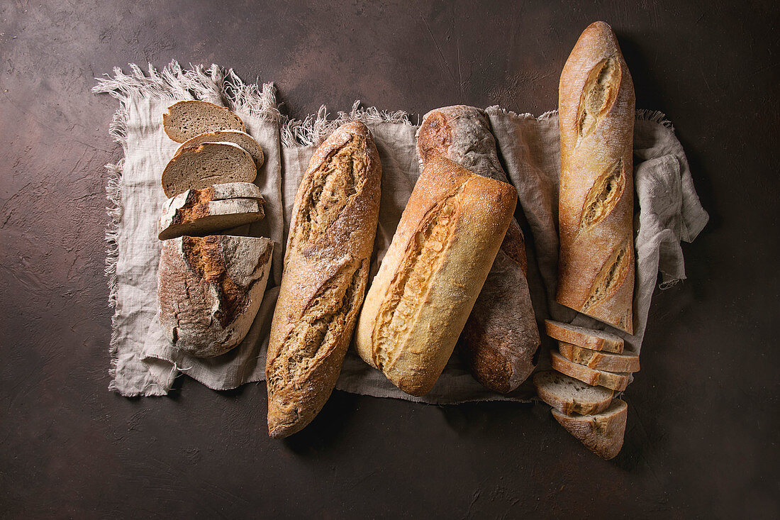 Variety of loafs fresh baked artisan rye, white and whole grain bread on linen cloth over dark brown texture background