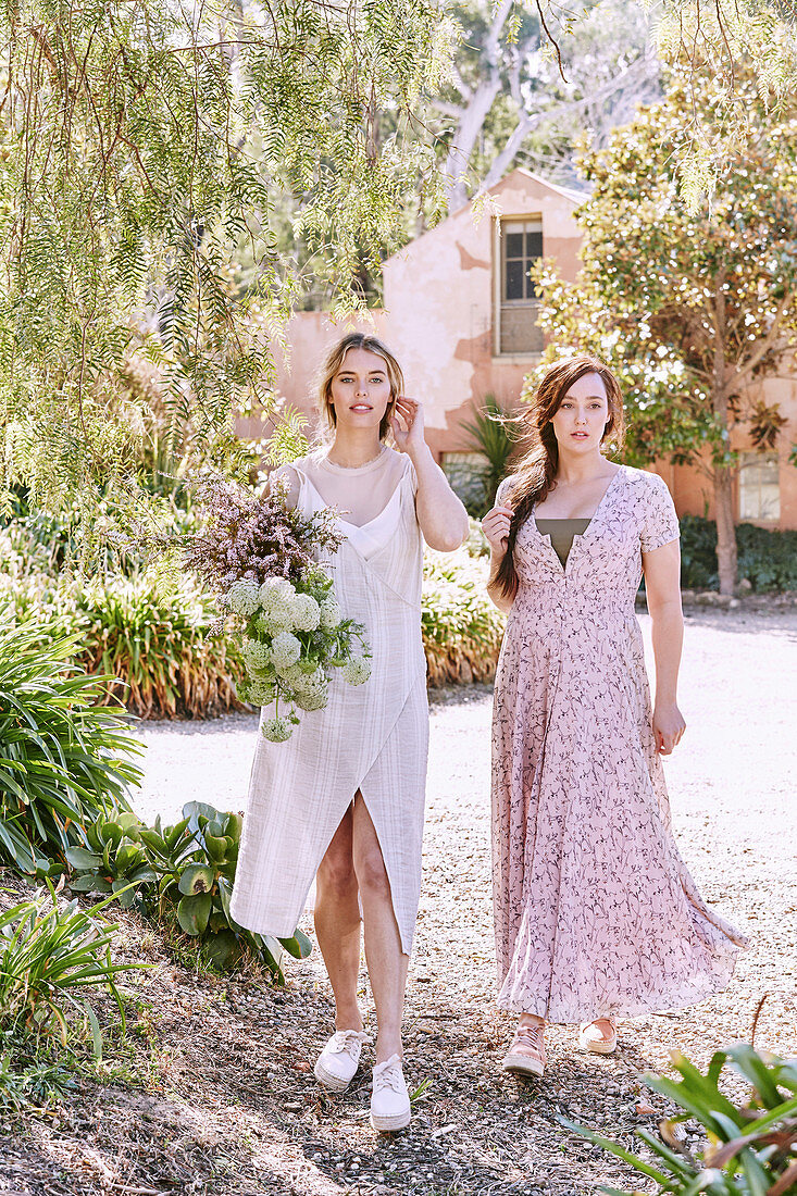 A blonde woman wearing a summer dress holding a bunch of flowers and a brunette woman wearing a delicate pink summer dress