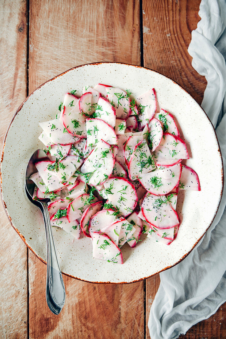 Radish salad with yogurt and poppy seed dressing in a white ceramic bowl on a wooden background from top view