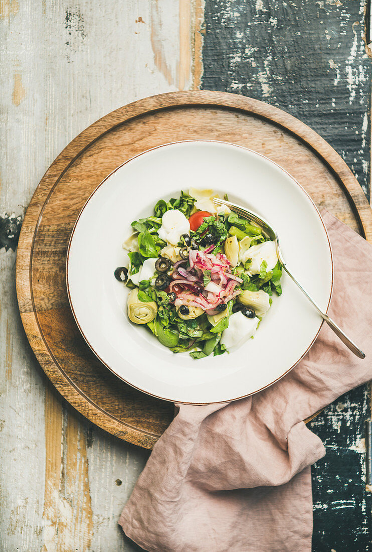 Flat-lay of fresh green summer salad with artichokes, olives, soft cheese and red onion in white plate over rustic wooden background