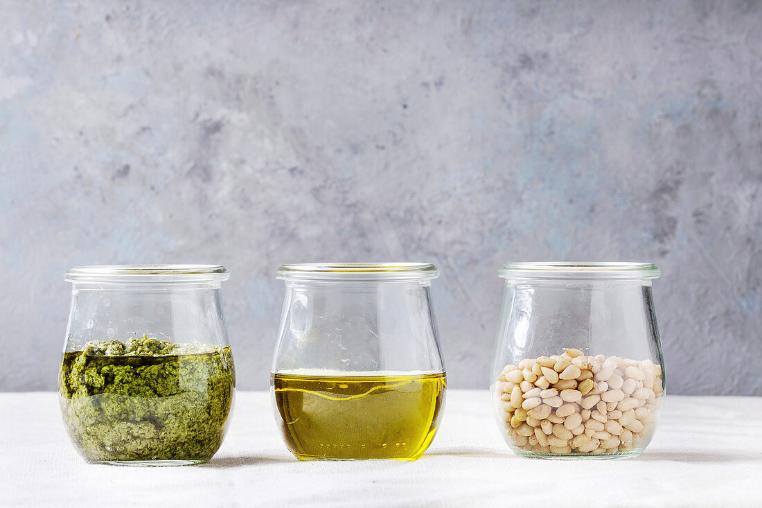 Glass jars with traditional Basil pesto sauce, olive oil, pine nuts on kitchen table with white linen tablecloth