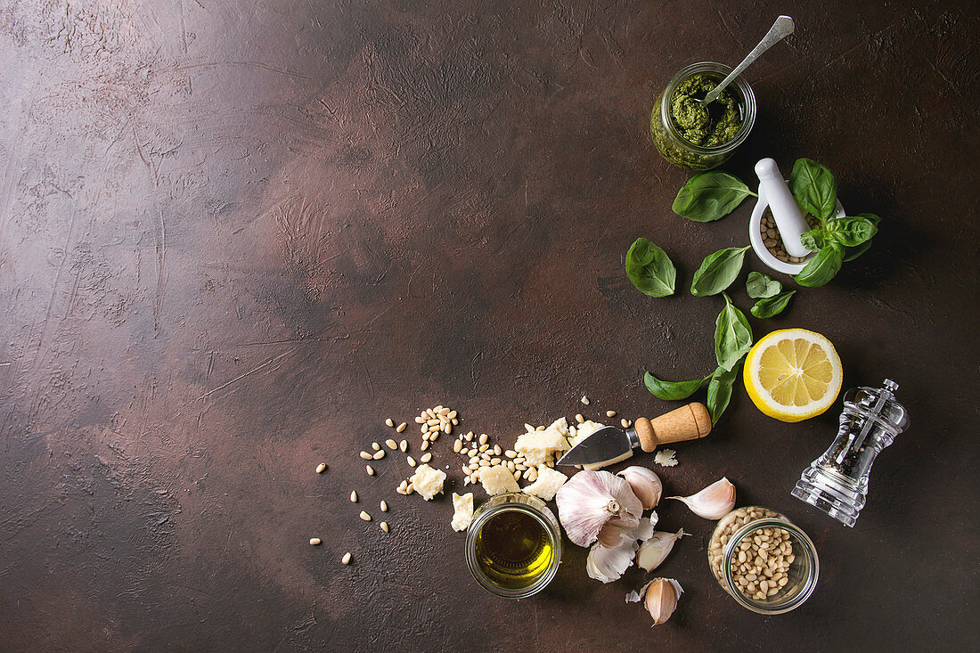 Traditional Basil pesto sauce in glass jar with ingredients above fresh basil, olive oil, parmesan cheese, garlic, pine nuts, lemon over dark texture background