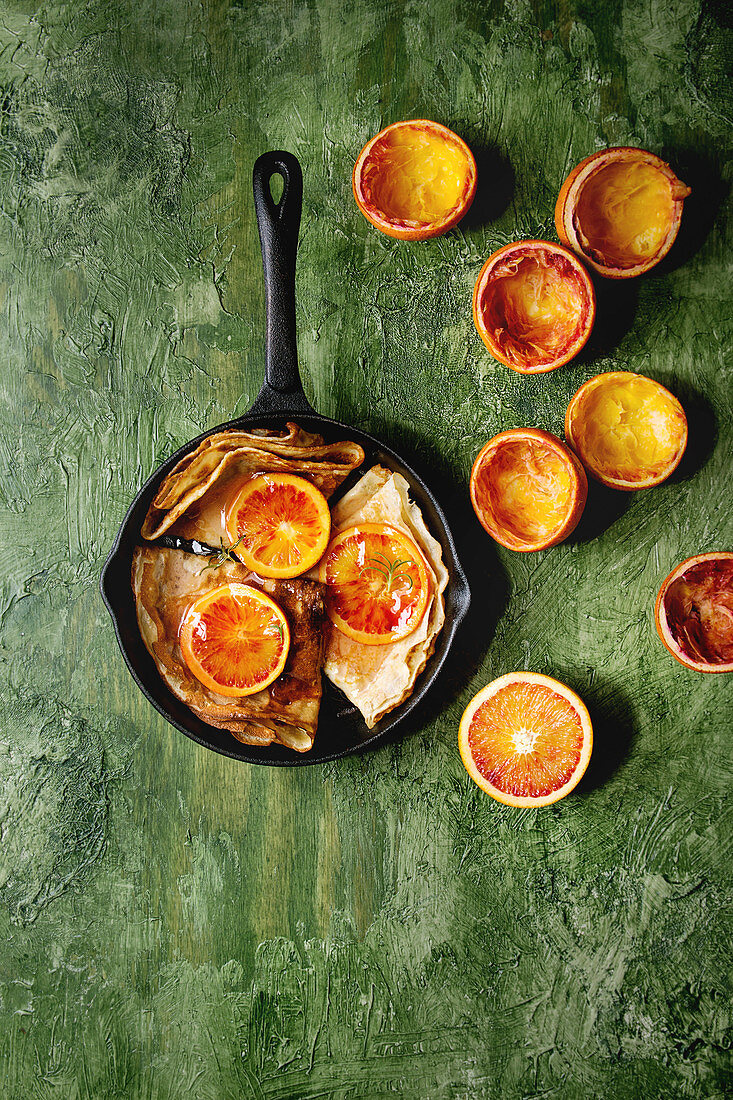 Homemade crepes pancakes served in cast-iron pan with bloody oranges and rosemary syrup with sliced sicilian red oranges over green texture background