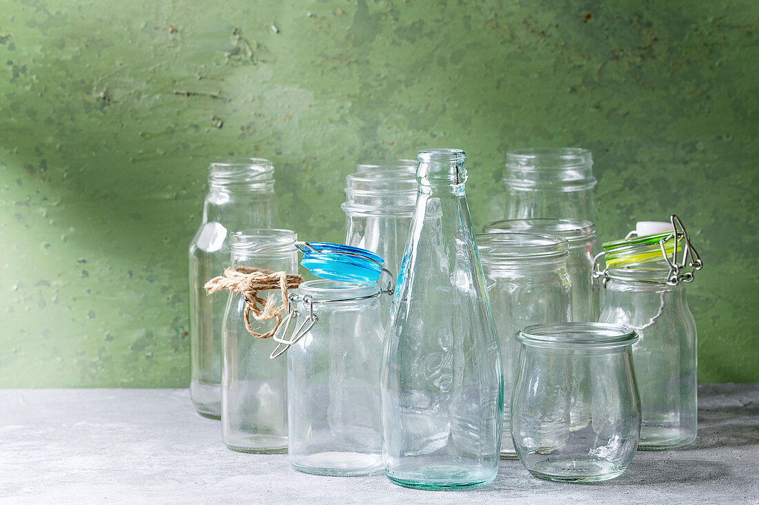 Variety of different shape empty opened glass bottles with and without lids standing on grey table with green wall as background