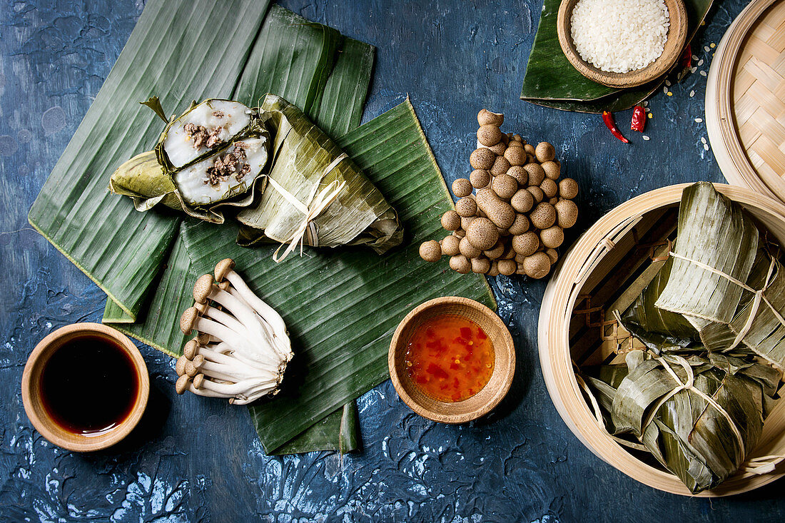 Asian rice piramidal steamed dumplings from rice tapioca flour with meat filling in banana leaves served in bamboo steamer. Ingredients and sauces above over blue texture background