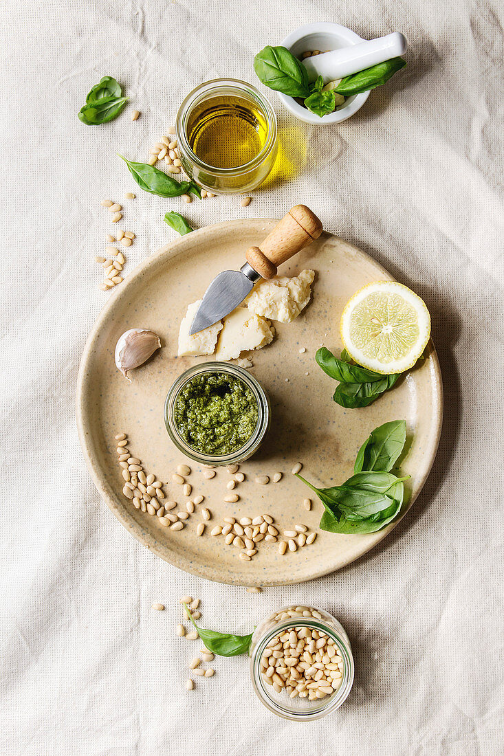 Traditional Basil pesto sauce in glass jar with ingredients above fresh basil, olive oil, parmesan cheese, garlic, pine nuts, lemon on ceramic plate over white linen cloth background