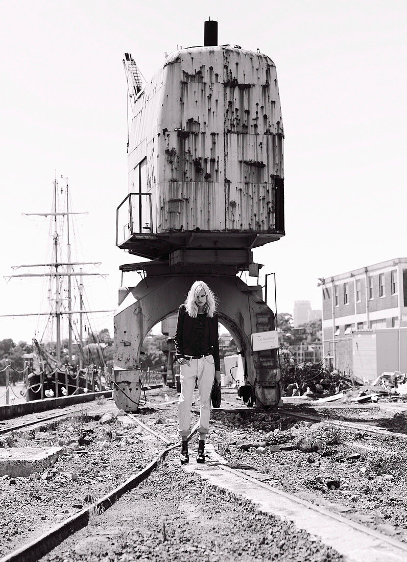 A blonde woman wearing a black jacket and white jeans on an industrial site (black-and-white shot)