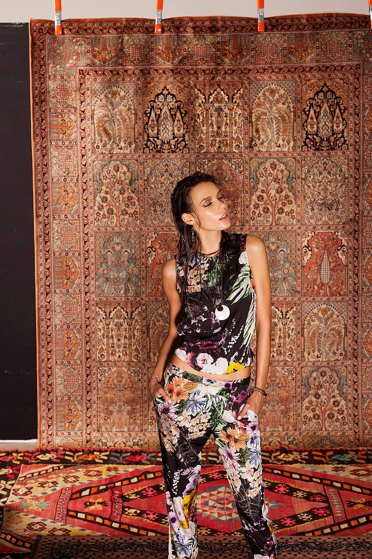 A young woman wearing a floral patterned top and trousers standing in front of a hanging rug