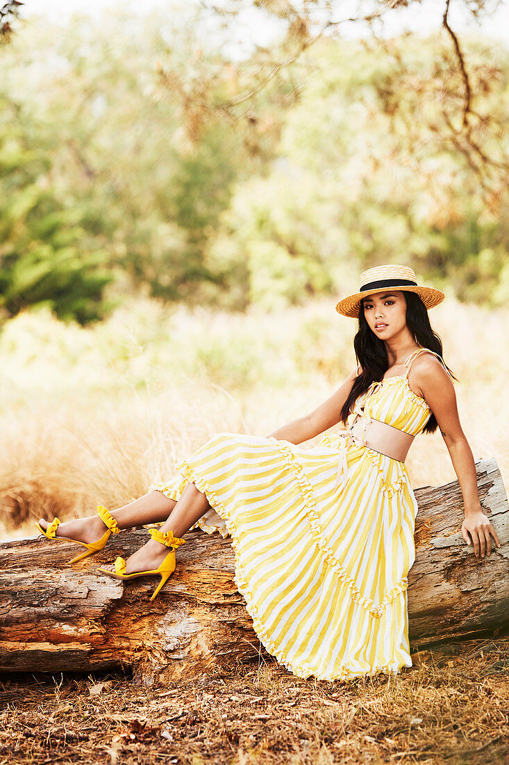A young dark-haired woman wearing a hat and a yellow-and-white striped summer dress sitting on a log