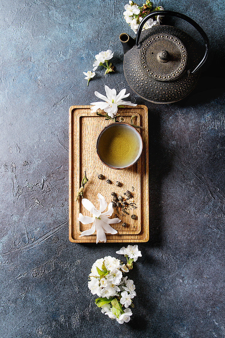 Traditional ceramic cup of hot green tea on wooden board with black iron teapot, spring flowers white magnolia and cherry blooming branches