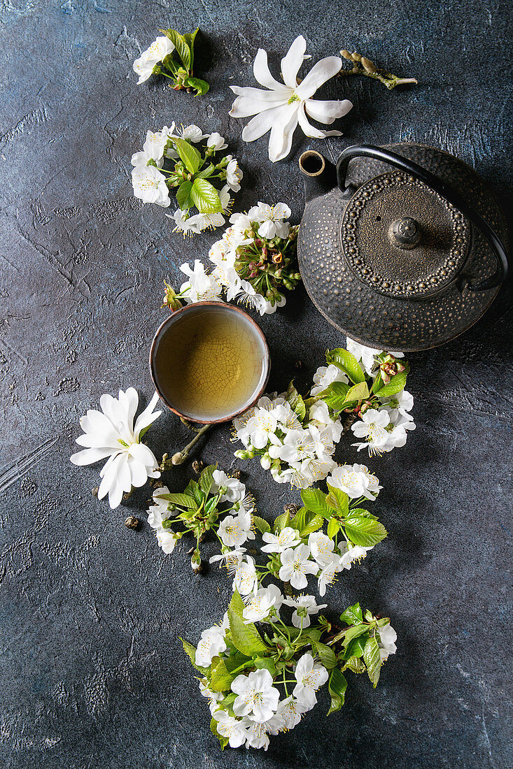 Traditional ceramic cup of hot green tea with black iron teapot, spring flowers white magnolia and cherry blooming branches