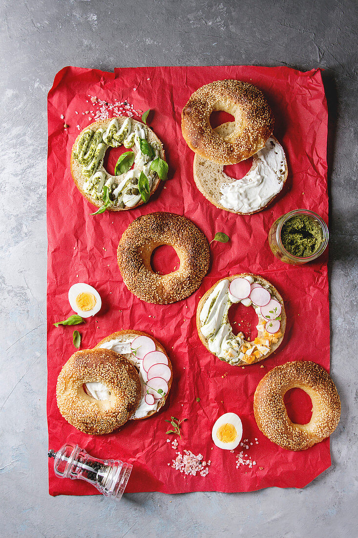 Variety of homemade bagels with sesame seeds, cream cheese, pesto sauce, eggs, radish, herbs served on red crumpled paper