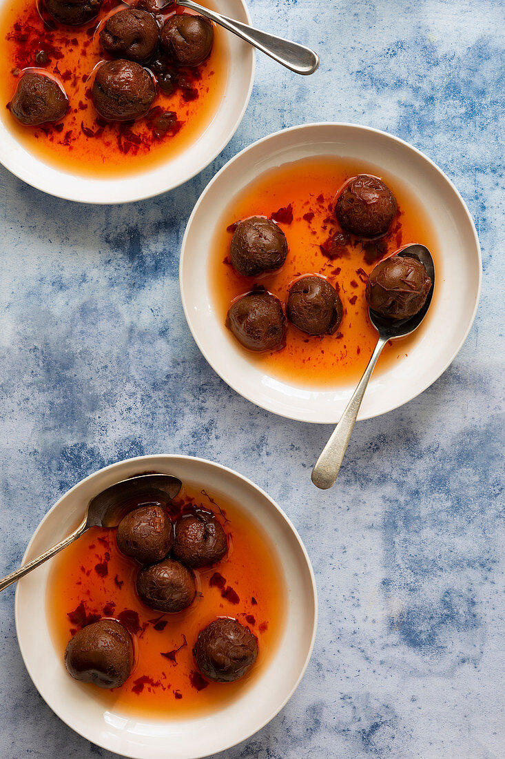 Three bowls of stewed red plums in syrup with spoons on a blue textured background