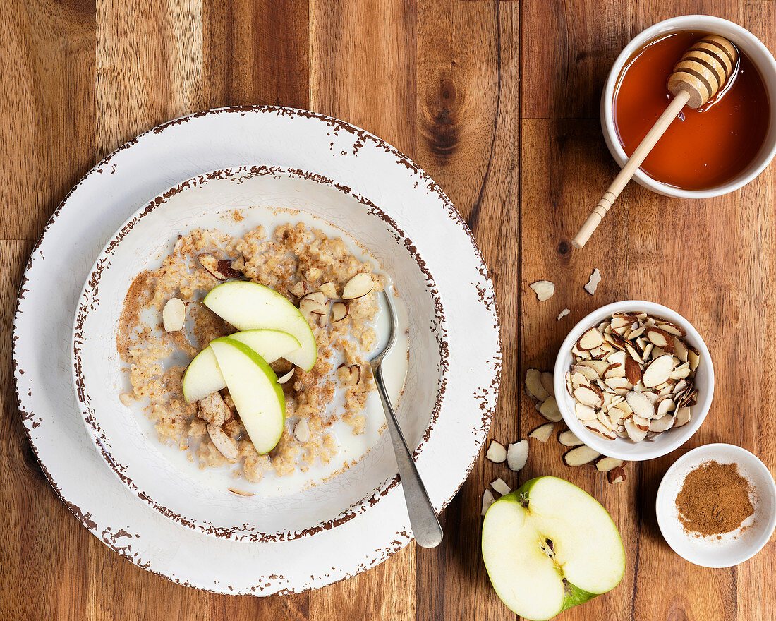 A bowl of organic oatmeal porridge with sliced apple on top and bowls of flaked almonds and cinnamon