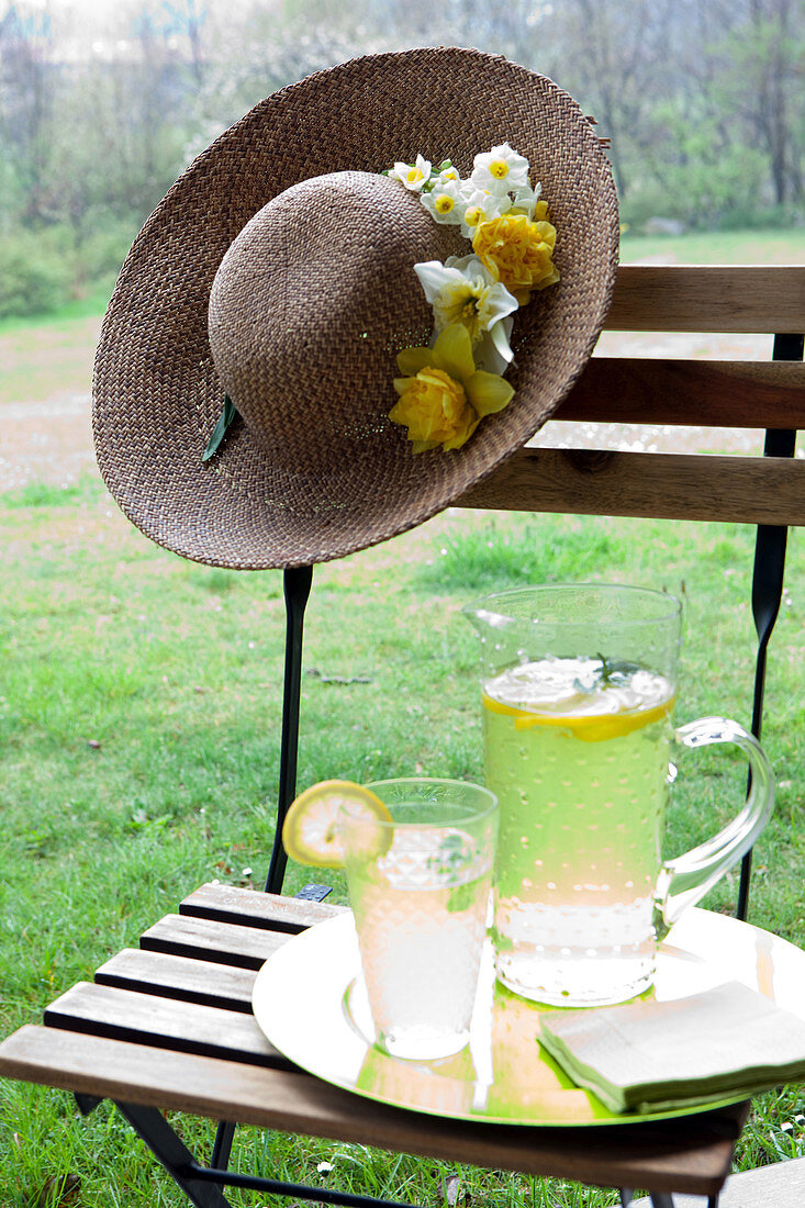 Jug and glass of lemonade and summer hat decorated with flowers on garden chair