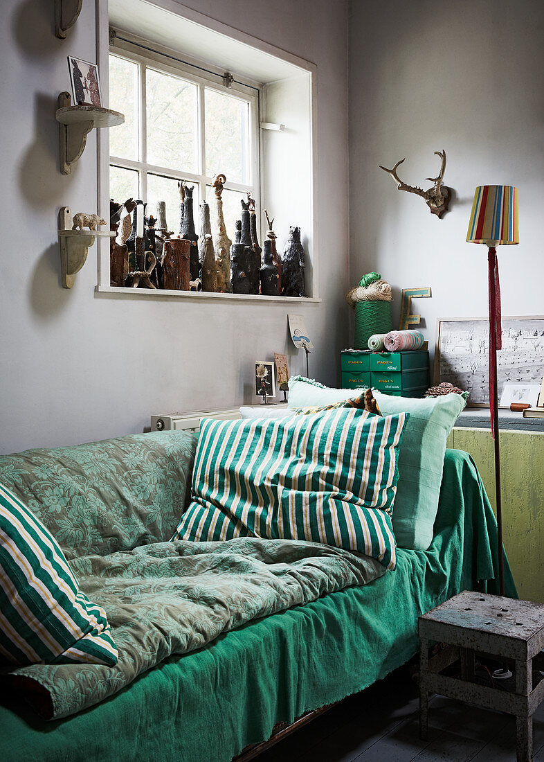 Green blankets and cushions on sofa below window with ornaments on windowsill