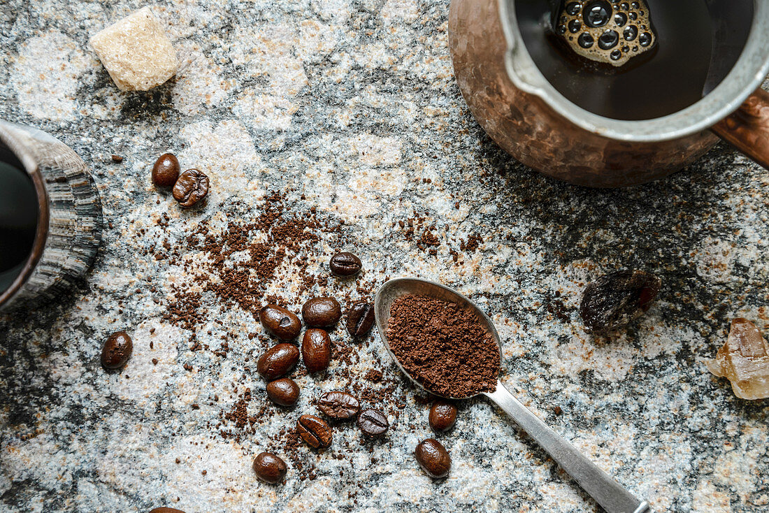 Top down view of scattered coffee beans, ground coffee in a spoon, brewed coffee in jezva on a granite surface