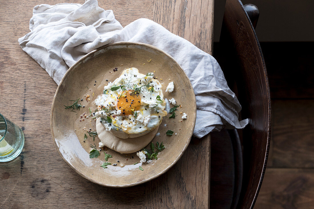 Fried eggs, feta on pita bread with fresh herbs on a wooden table and a chair