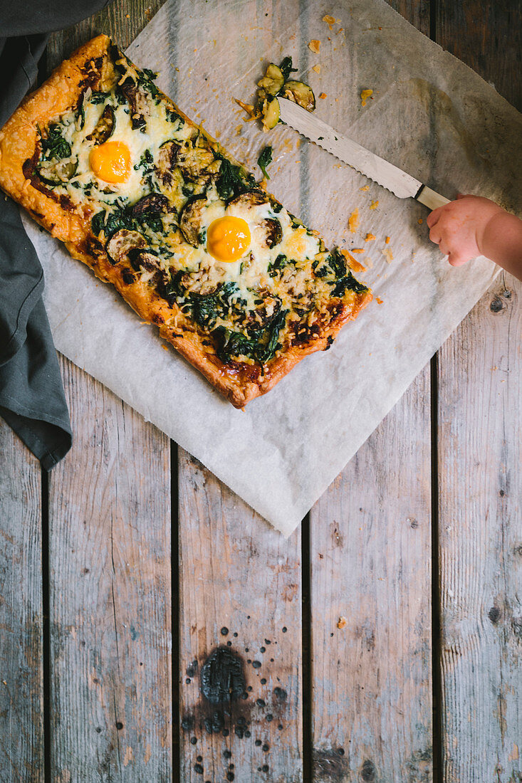 Pizza with spinach, fried eggs, courgette, melted cheese and a small child hand