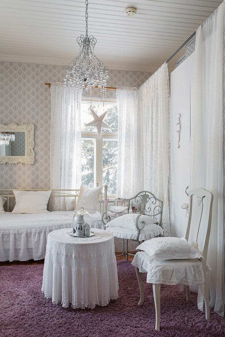 Runder Tisch mit bodenlanger Tischdecke, Stühle und Metallbett in weißem Zimmer