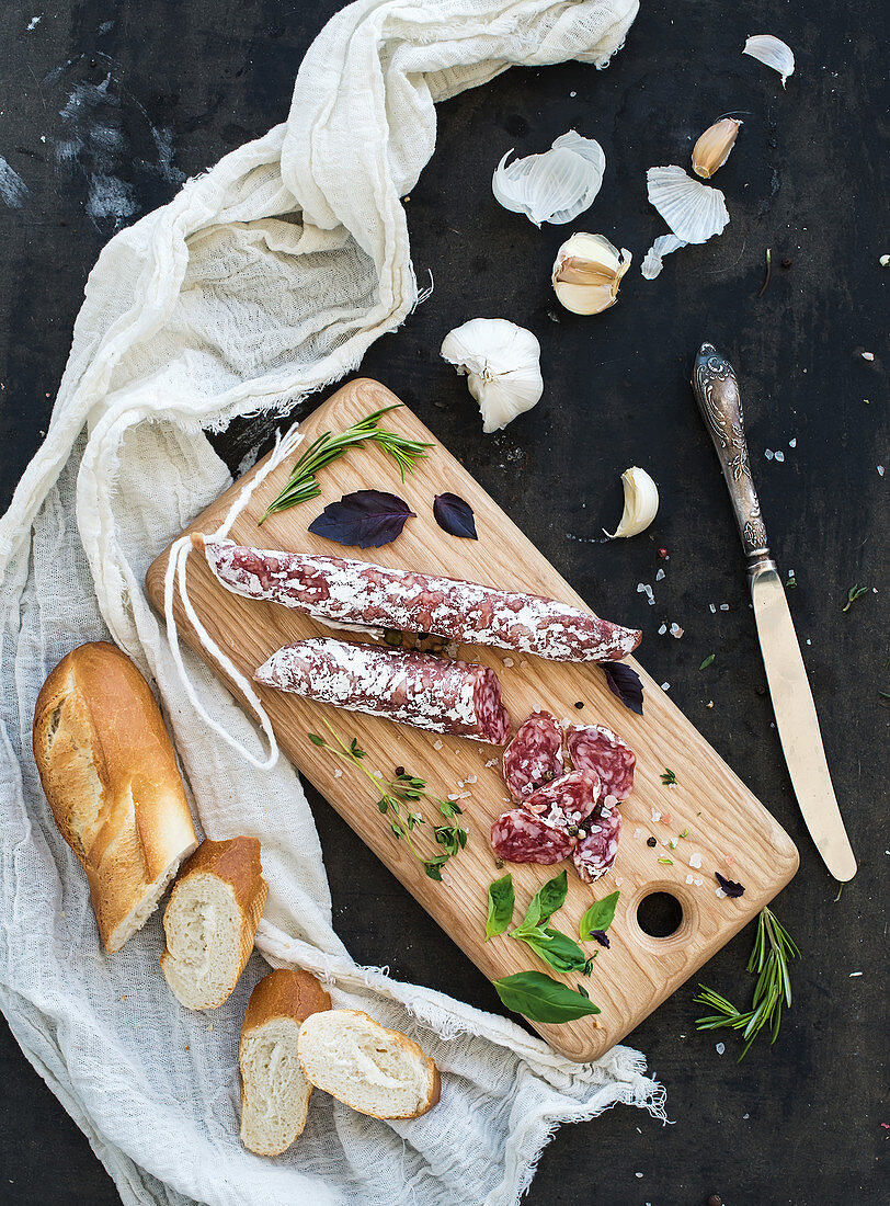 Salami, garlic, baguette and herbs on rustic wooden board
