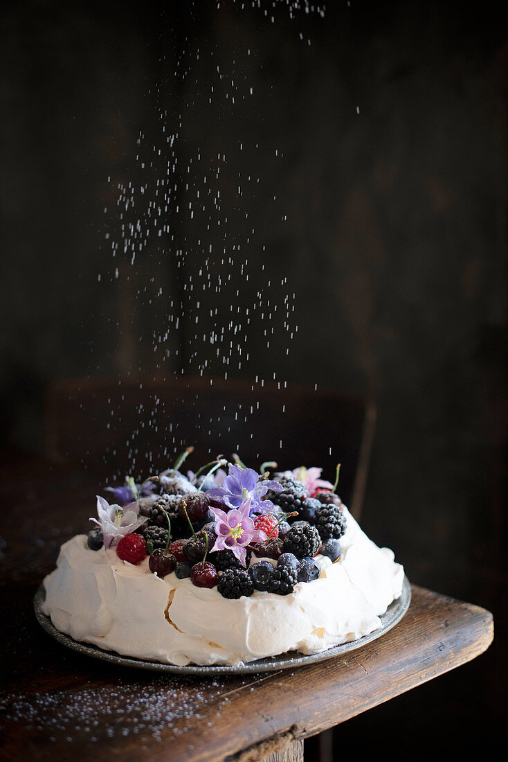 Icing sugar being sprinkled over a pavlova with berries and cherries