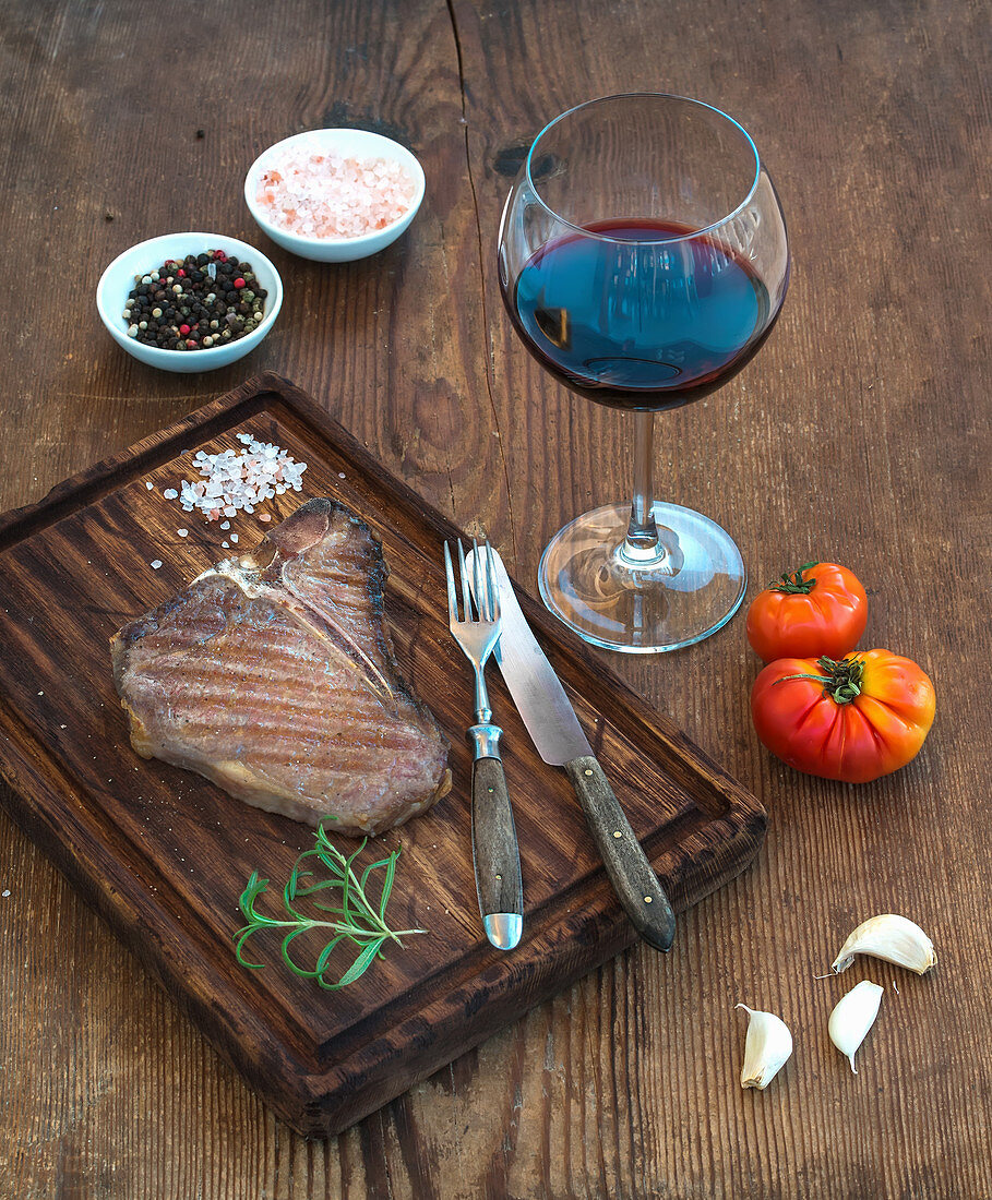 Gegrilltes T-Bone-Steak mit Knoblauchzehen, Tomaten, Rosmarin, Gewürzen und Rotweinglas