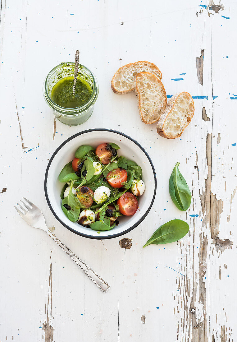 Caprese salad and chiabatta slices. Cherry-tomatoes, baby spinach and mozzarella in metal bowl with pesto dressing on rustic white wooden backdrop