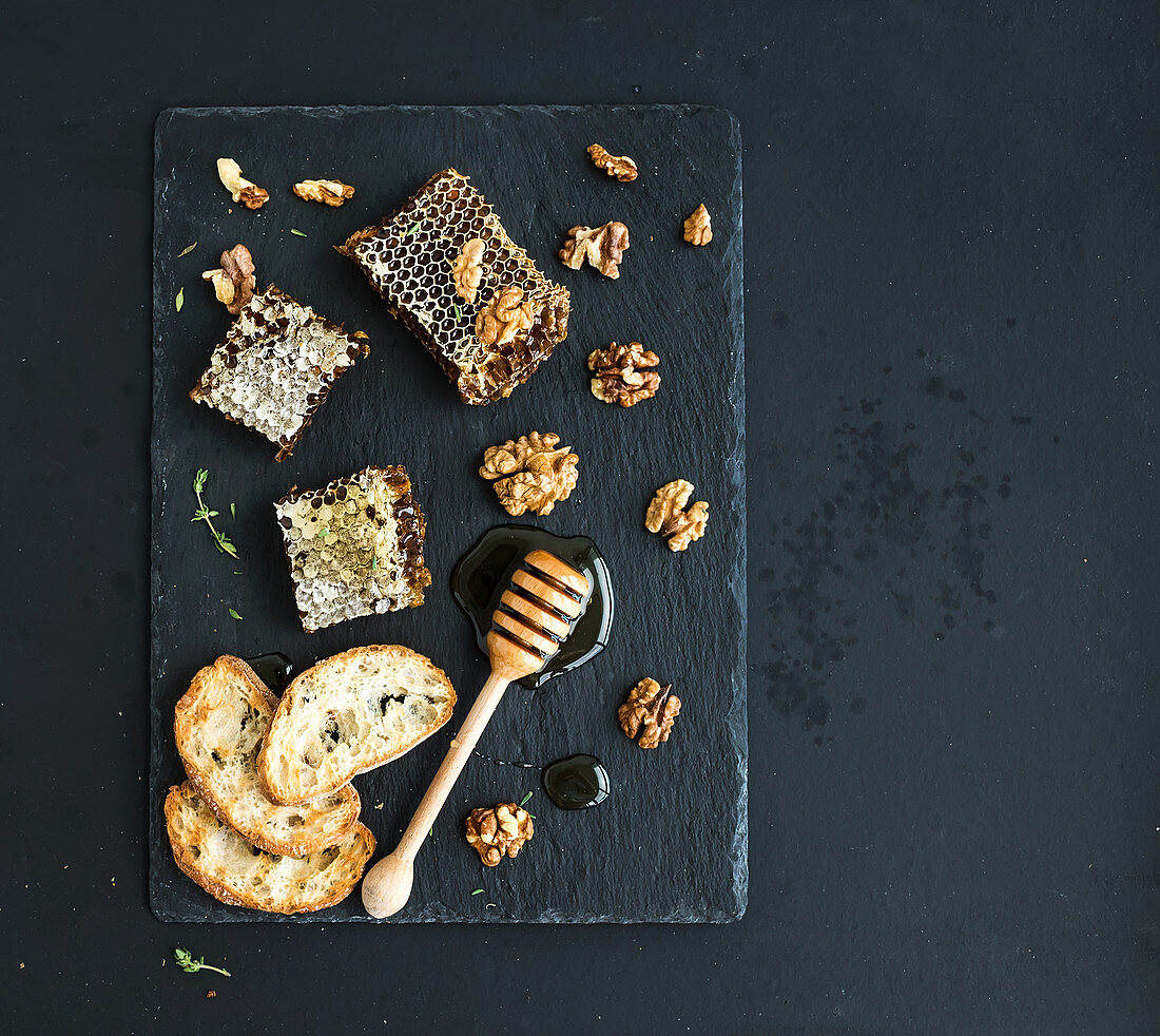 Honeycomb, walnuts, bread slices and honey dipper on black slate tray over grunge dark backdrop