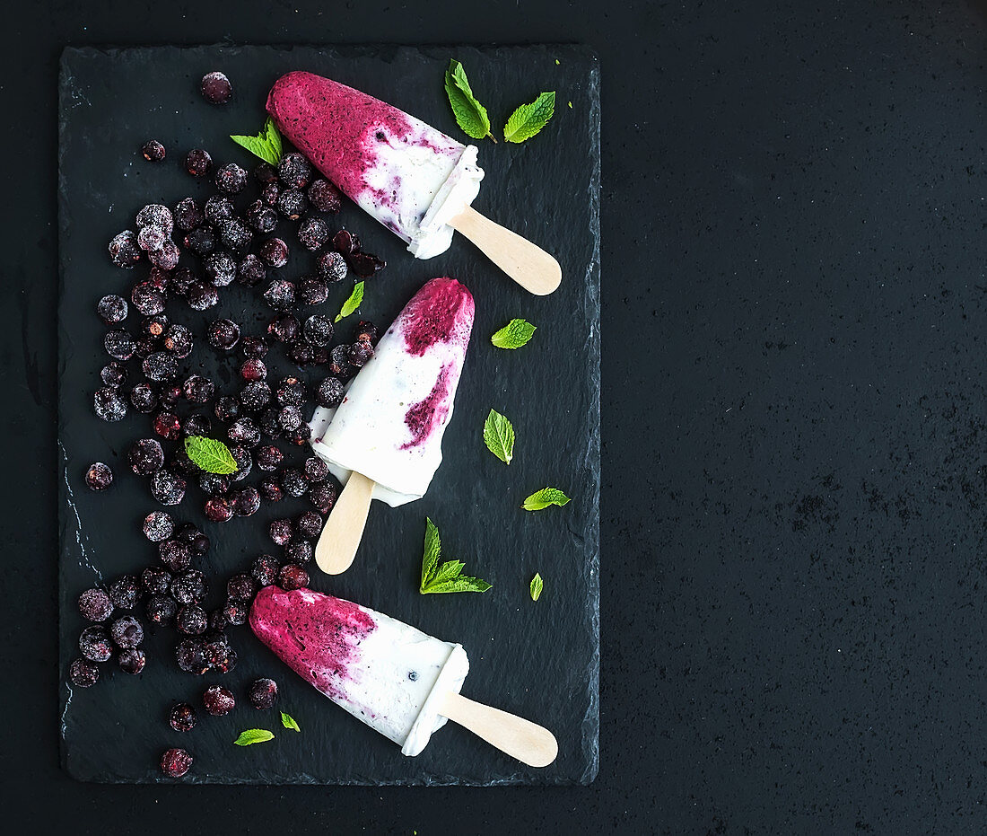 Black-currant and cream ice-creams or popsicles with frozen black-currant and mint on black slate tray over dark grunge backdrop