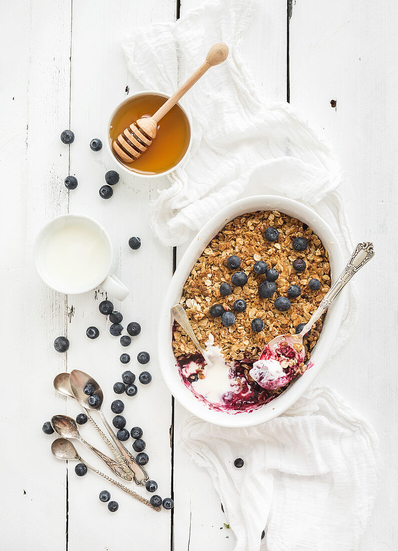 Healthy breakfast. Oat granola berry crumble with fresh blueberries, yogurt and honey in ceramic cooking dish over white rustic backdrop