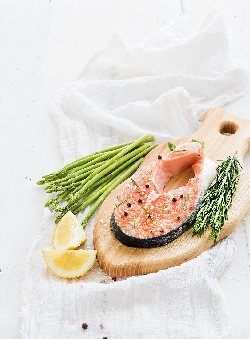 Raw salmon steak with asparagus, lemon, spices and rosemary on rustic wooden chopping board over white backdrop