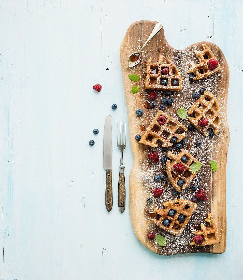 Soft Belgian waffles with berries, honey and mint on rustic wooden serving board over light blue background