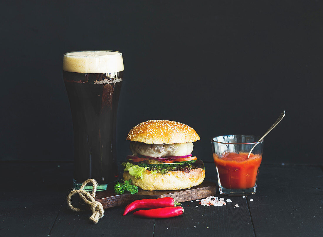 Fresh homemade burger on wooden serving board with spicy tomato sauce, sea salt, herbs and glass of dark beer over black background