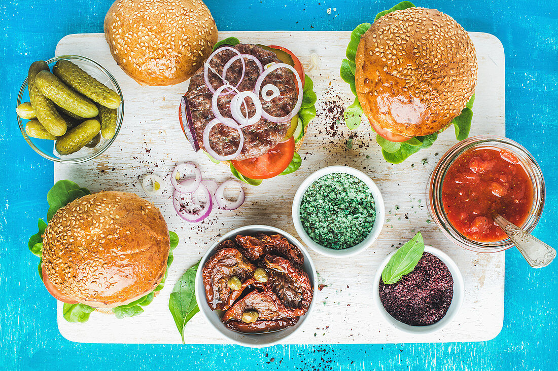 Hausgemachte Beefburger mit Zwiebelringen, Essiggurken, Gewürzen, getrockneten Tomaten und Tomatensauce