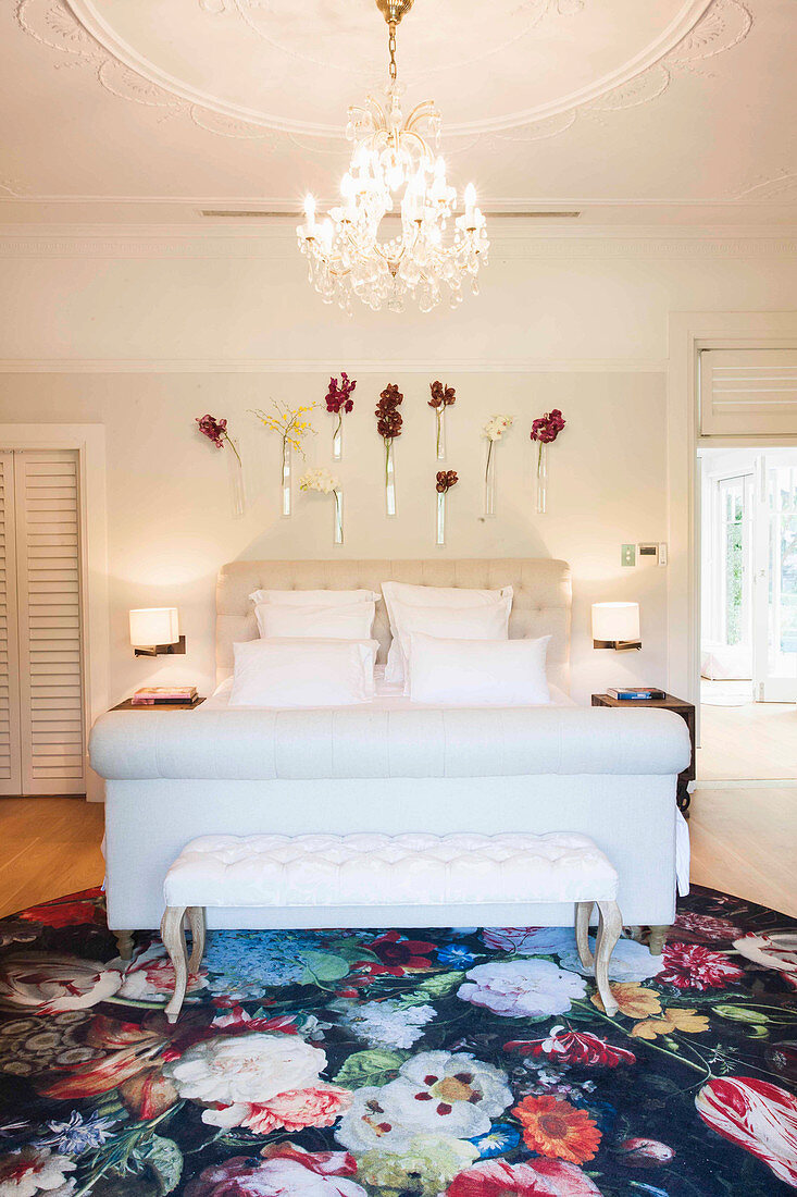 Elegant bedroom with white double bed, clothes bench, chandelier and flowers on the wall
