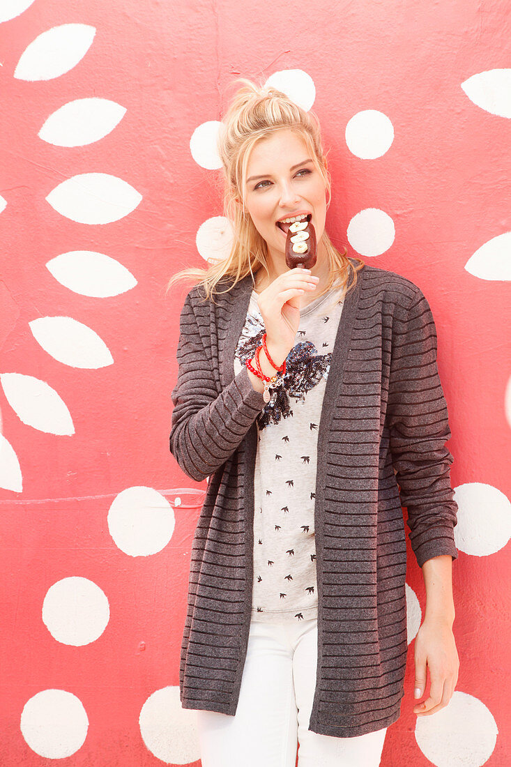 A blonde woman wearing a top and a cardigan eating an ice cream