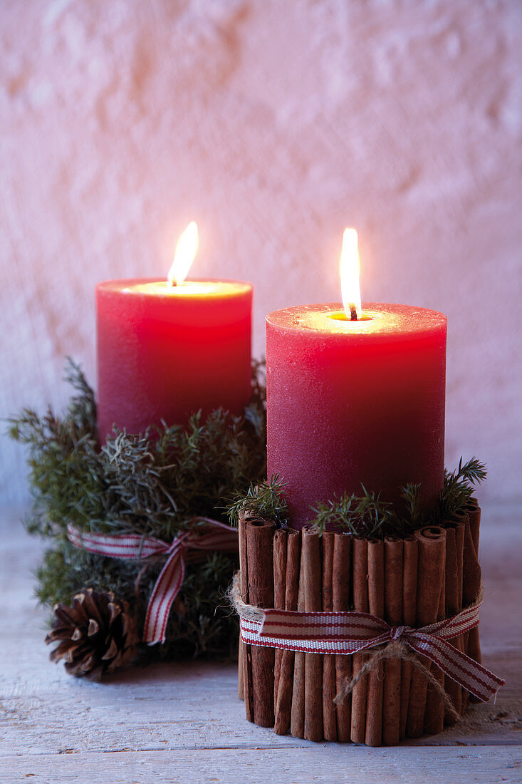 Two red pillar candles festively decorated with moss, pine cones and cinnamon sticks