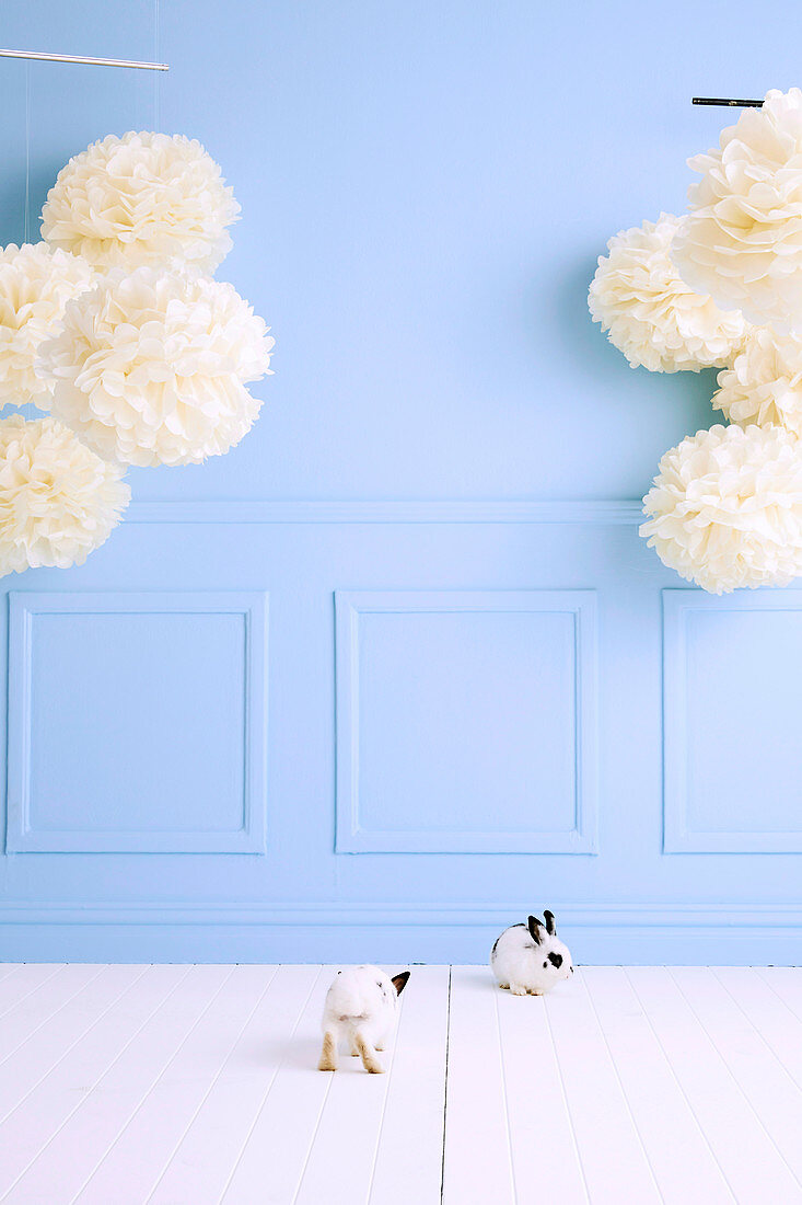 Two bunnies in the room with a light blue wall and huge paper flowers