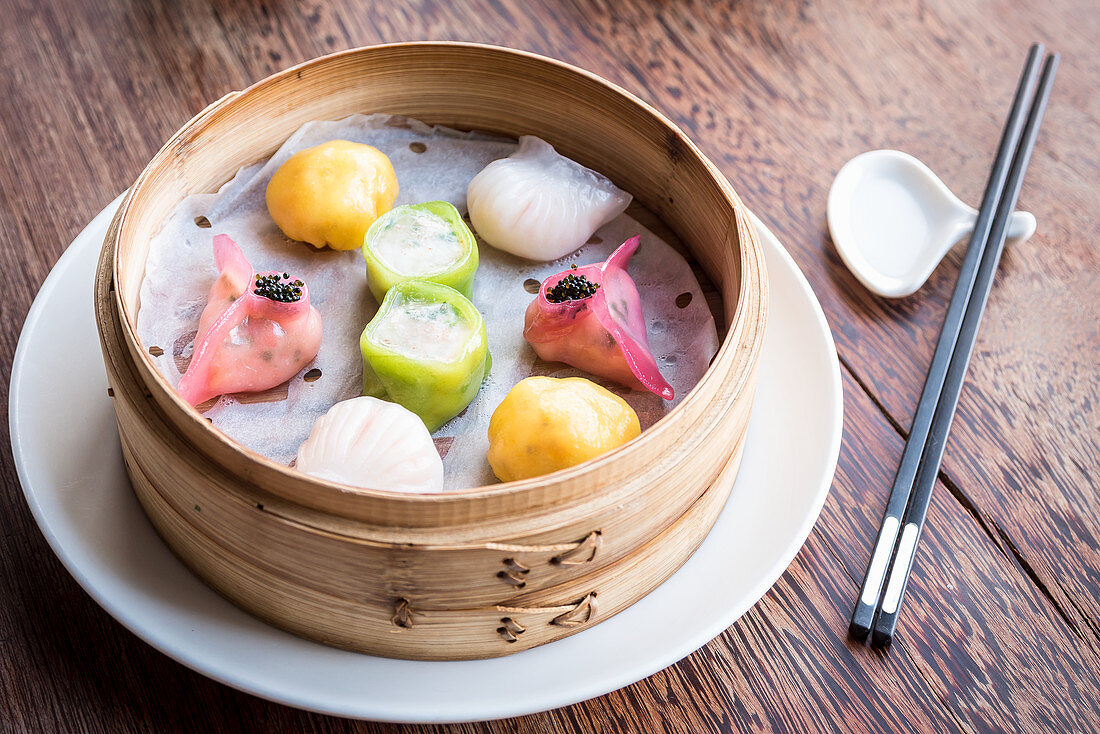 Assorted and colourful dim sum dumplings in a bamboo steamer on a wooden table with chopsticks