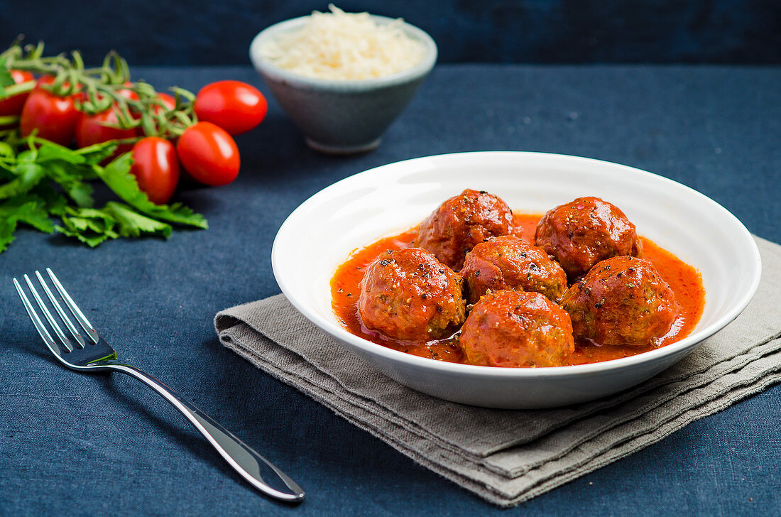 Plate of meatballs in tomato sauce with black pepper on a blue tablecloth with fork and ingredients in the background