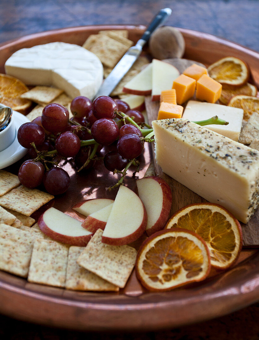 Käseplatte mit Brie, Cheddar und Kräuterkäse, Crackern, getrockneten Orangen, Trauben und Äpfeln auf Kupfertablett