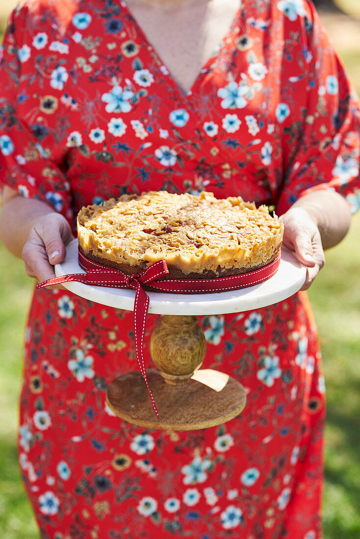 Frau hält Mandel-Karamellkuchen mit eingelegten Kirschen auf Tortenständer
